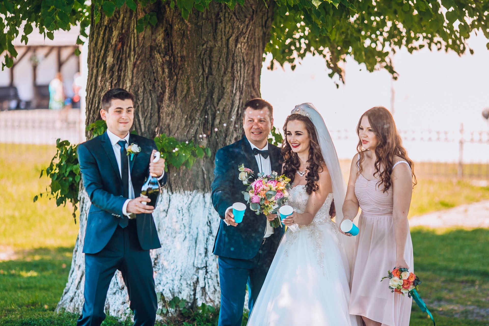 Newlyweds and their friends open champagne bottle standing in the park. Before wedding ceremony friends having fun and drink champagne. Funny wedding moment of newlyweds and bridesmaids and groomsmen.
